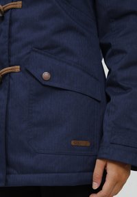 Marinblå jacka tillverkad av strukturerad tyg med en fiskbensmönster, utrustad med detaljer i brunt rep, en logoknapp och en sidoficka.