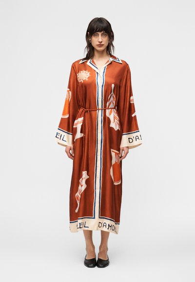 Femme portant une robe midi boutonnée couleur rouille avec des manches larges, des imprimés abstraits, une fine ceinture à la taille, et des chaussures plates noires.