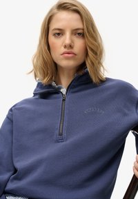 Granatowy półgolf zapinany na zamek z miękkiej tkaniny z wyszywanym logo "Superdry". Posiada luźny krój i szczegóły z ribbingu przy mankietach.