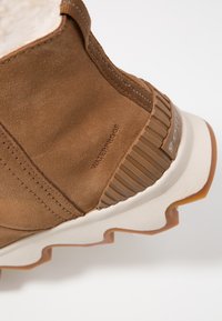 Wodoodobny brązowy zamszowy botek za kostkę o fakturowanej powierzchni, białej podeszwie oraz żebrowanych akcentach na pięcie dla lepszej przyczepności i stylu.