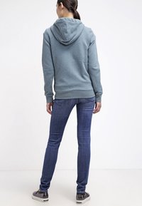 Felpa grigia con cappuccio regolabile, orlo e polsini a coste; abbinata a jeans blu scuro e sneaker grigie. Vista posteriore.
