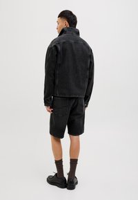 Zwarte spijkerjacket met hoge kraag, gecombineerd met zwarte denimbroekjes. Model draagt zwarte schoenen en bruine sokken, staande tegen een effen achtergrond.