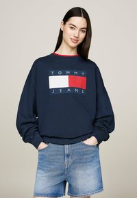 Felpa blu navy con un rettangolo logo bianco e rosso e testo "TOMMY JEANS". Collo a giro e vestibilità comoda. Pantaloni shorts di denim con tasche.