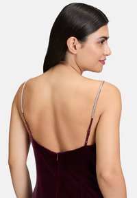 Robe en velours bordeaux avec un dos nu et des bretelles fines. Les bretelles sont ornées. Texture lisse et design ajusté avec une fermeture éclair.