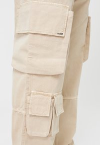 Beżowe spodnie cargo wykonane z wytrzymałego materiału, z wieloma dużymi kieszeniami, kieszenią na zamek i widocznymi detalami szycia.
