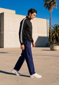 Ung man i svart jacka med vita ränder, marinblå byxor och vita sneakers som promenerar utomhus nära en modern betongbyggnad och palmer.