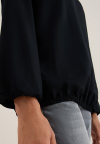 Close-up van een persoon in een zwart langemouwen shirt met elastische manchetten en lichtgrijze broeken, waarbij de taille en handen zichtbaar zijn.