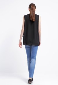 Svart ärmlös topp med lös passform, matchad med blå skinny jeans och svarta skor, sedd bakifrån mot en neutral bakgrund.