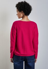 Maglione rosa loose-fit con scollatura ampia, tessuto testurizzato e orlo a coste. Abbinato a jeans in denim blu scuro.