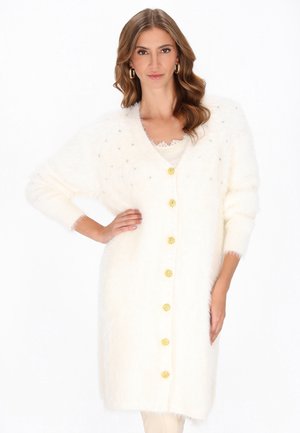 Eine Frau mit welligem Haar, die einen langen weißen, flauschigen Cardigan mit goldenen Knöpfen, Strassdetails und goldenen Creolen trägt, vor einem weißen Hintergrund.