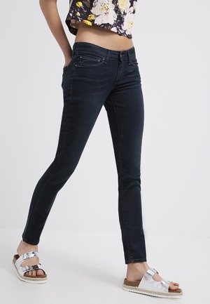 Personne portant un jean skinny foncé, un top court fleuri et des sandales à boucle argentée, debout les mains dans les poches.