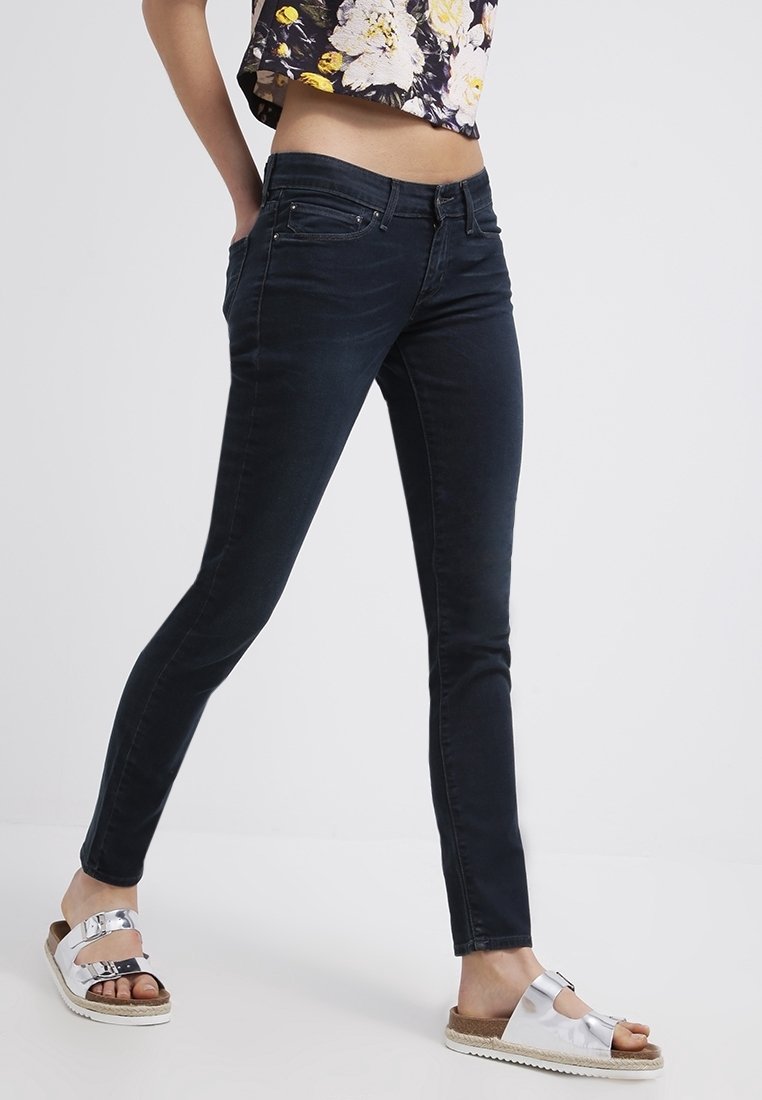Personne portant un jean skinny foncé, un top court fleuri et des sandales à boucle argentée, debout les mains dans les poches.