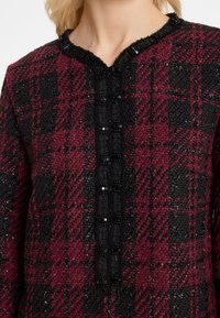 Rot-schwarze karierte Tunika aus strukturiertem Stoff, mit Rundhalsausschnitt und schwarzen Knöpfen entlang der Vorderseite. Subtile Glitzerelemente sind sichtbar.