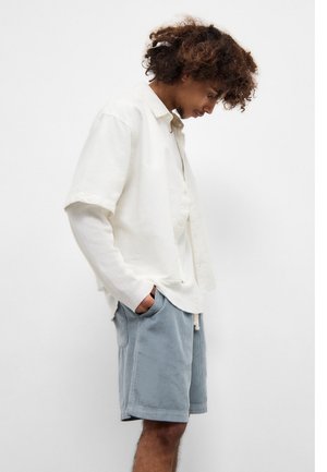 Hvid kortærmet skjorte med knapper over en lysegrå ribbet t-shirt, sat sammen med lysegrå fløjlsshorts. Simpelt design, afslappet pasform.