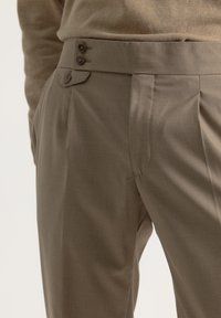 Pantalones a medida en una tela marrón apagado, con frente plano, pliegues marcados, dos botones en la cintura y una textura suave.