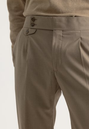Pantalones a medida en una tela marrón apagado, con frente plano, pliegues marcados, dos botones en la cintura y una textura suave.