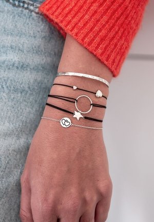 Schwarze Kordelarmbänder mit silbernen Akzenten, die ein Herz, einen Stern und kreisförmige Formen enthalten. Ein Metallarmband mit eingraviertem Text ist ebenfalls enthalten.