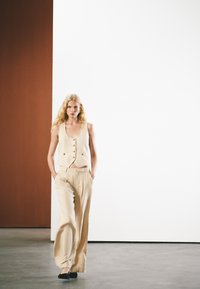 Beige linnen vest met knopen, gecombineerd met wijde beige linnen broeken. Het model staat zelfverzekerd en toont een minimalistisch ontwerp.