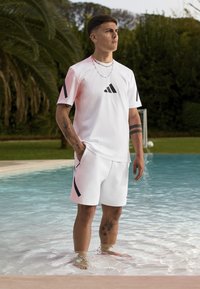 Hvit atletisk t-skjorte med svarte detaljer og logo, kombinert med hvite shorts med svarte side-detaljer. Modell som står i grunt vann.