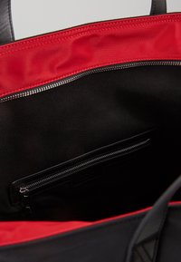 Sac fourre-tout noir ouvert montrant une doublure intérieure rouge et une poche intérieure zippée avec des anses en tissu noir et en cuir.