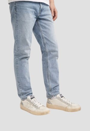 Persona che indossa jeans azzurri chiari e sneakers bianche con design a stella, in piedi su uno sfondo grigio chiaro uniforme.