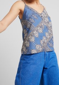 Top con pattern blu, spalline sottili regolabili e dettaglio con bottoni, con motivi decorativi a paisley e forme geometriche. Indossato con pantaloni in denim.