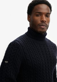 Dunkelblauer, kabelgestrickter Rollkragenpullover aus dickem Garn, mit einem gerippten Kragen und einem kleinen Markenzeichen auf dem linken Ärmel.