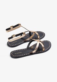 Black sandals with metallic gold straps, featuring a flat design, buckle closure, and studded detailing along the edge of the sole.