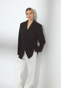 Melna žakete ar atlocītiem uzlikām, kombinēta ar brīvām baltām biksēm, fiksēta uz balta fona.