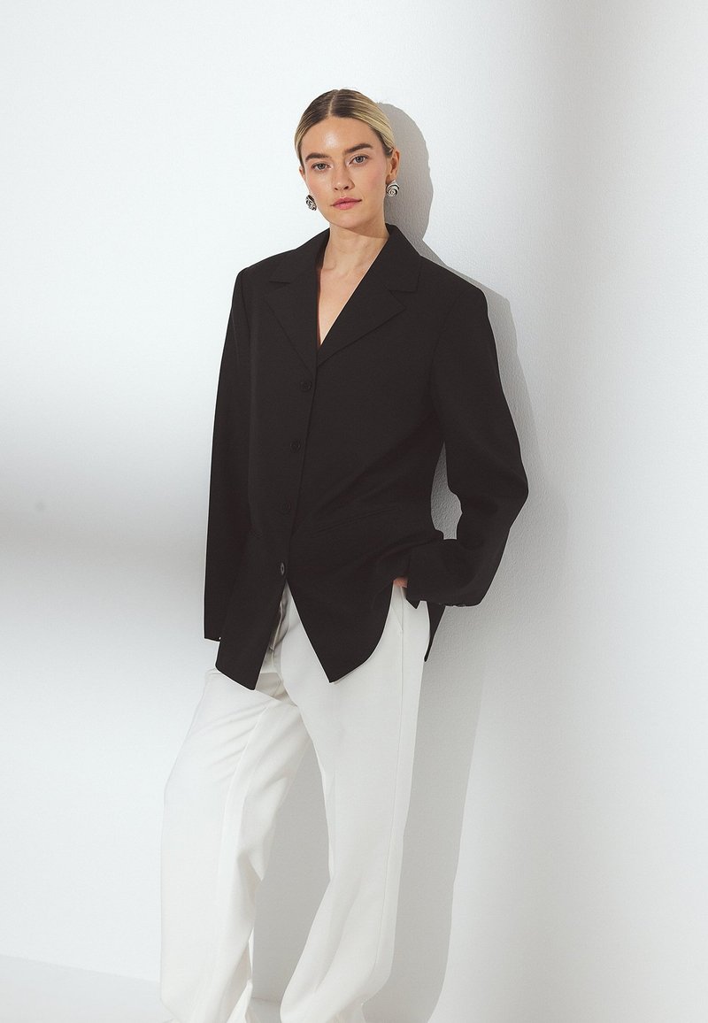 Melna žakete ar atlocītiem uzlikām, kombinēta ar brīvām baltām biksēm, fiksēta uz balta fona.