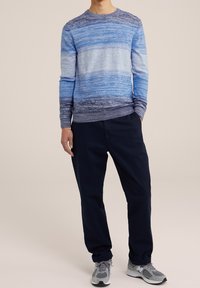 Modrý proužkovaný svetr v různých odstínech modré, doplněný tmavě modrými kalhotami. Textura působí měkce a pleteně. Šedé sportovní boty.