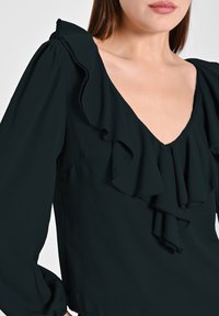 Blusa verde scuro con scollatura a V profonda, dotata di volant a strati, maniche lunghe a palloncino e tessuto morbido e leggero per un aspetto fluido.