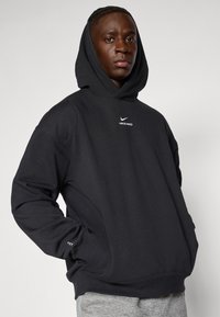 Černá mikina Nike Pro s klokaní kapsou, žebrovanými manžetami a malým bílým logem na prsou. Měkká tkanina s volným střihem.