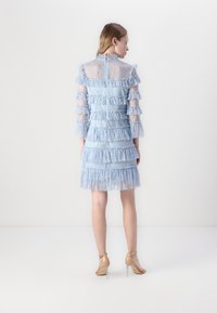 Abito in pizzo blu chiaro con volants a strati, lunghe maniche trasparenti e collo alto. Chiusura con cerniera sul retro e tacchi con cinturino alla caviglia.