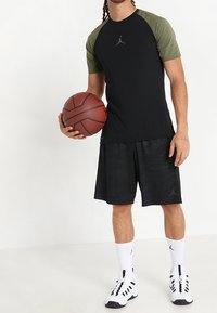 Svart kortärmad träningsskjorta med olivgröna raglanärmar, matchad med svarta mönstrade shorts, vita strumpor och skor, hållande en basketboll.
