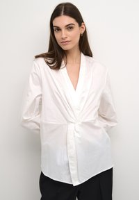 Blusa bianca a maniche lunghe con scollo a V profondo, frontale avvolgente e dettaglio annodato in vita. Realizzata in un tessuto liscio e leggero.