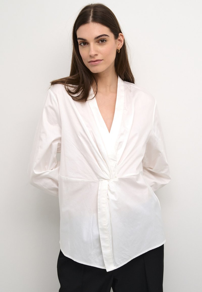 Blusa bianca a maniche lunghe con scollo a V profondo, frontale avvolgente e dettaglio annodato in vita. Realizzata in un tessuto liscio e leggero.