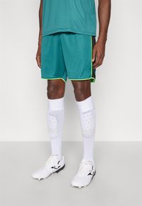 Tirkīza krāsas sporta šorti ar neona zaļiem akcentiem, kopā ar baltām zeķēm un melni baltām futbola kurpēm. Spēlētājs valkā kāju aizsargus zem zeķēm.