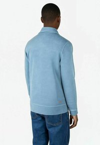 Mann, der sich abwendet, trägt ein hellblaues Langarmshirt und dunkelblaue Jeans vor einem schlichten weißen Hintergrund.