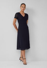 Robe bleu marine en tissu texturé, avec des manches courtes, un décolleté en V, et une jupe mi-longue avec une taille cintrée. Talons aiguilles noirs à lanières.