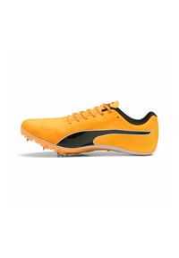 Zapatilla deportiva amarilla con detalles en negro, material sintético suave, seis pinchos metálicos en la suela y cordones naranjas.
