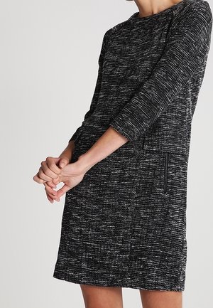 Kobieta ubrana w czarno-białą dzianinową sukienkę o fakturze, z rękawami do połowy przedramienia, z rękami złączonymi przed sobą, widoczna od szyi do połowy ud.