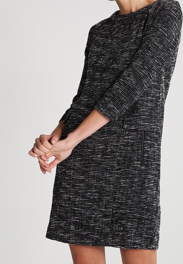 Femme portant une robe en maille texturée noire et blanche à manches trois-quarts, les mains jointes devant, vue du cou jusqu'à mi-cuisse.