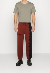 Braune Batik-Sweatpants mit schwarzen Akzenten, verstellbarem Bund und besticktem Logo, kombiniert mit einem schlichten grauen T-Shirt und schwarzen Stiefeln.