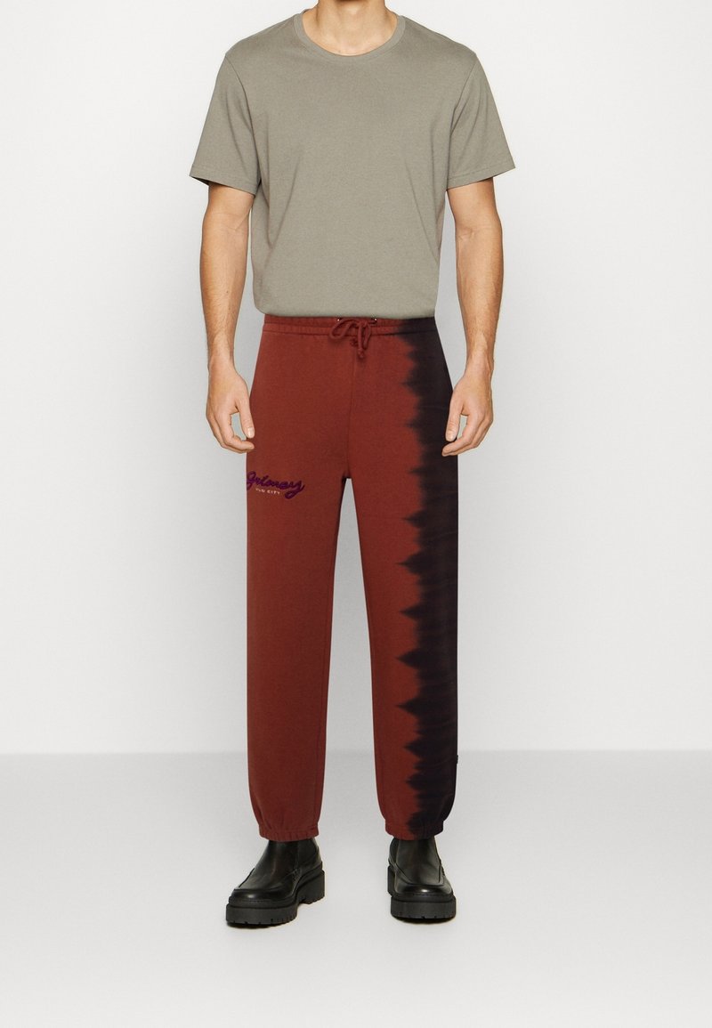 Braune Batik-Sweatpants mit schwarzen Akzenten, verstellbarem Bund und besticktem Logo, kombiniert mit einem schlichten grauen T-Shirt und schwarzen Stiefeln.