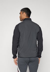 Chaqueta de hombre en gris oscuro con mangas negras, cuello alto y puños elásticos. Textura suave con diseño minimalista y sin logos visibles.