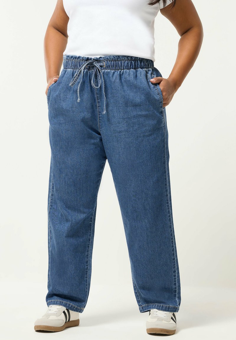 Osoba nosząca luźne niebieskie spodnie dżinsowe z elastycznym pasem i sznurkiem, białe sneakersy z czarnymi paskami, ręce w kieszeniach.