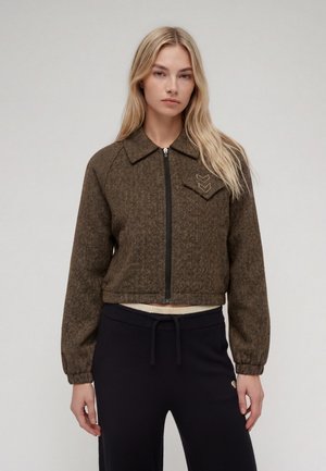 Femme aux longs cheveux blonds portant une veste zippée marron à motif chevrons et un pantalon noir sur un fond clair uni.