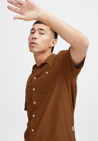 Camisa marrón de manga corta tipo botonadura en una tela suave, con un bolsillo en el pecho y botones blancos, con un corte relajado y cuello de camisa.