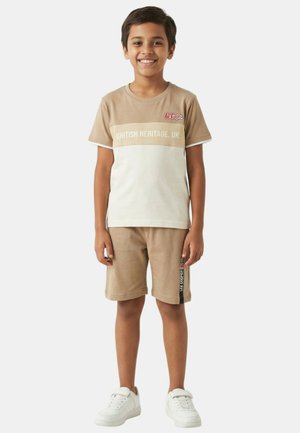 Un garçon souriant se tient debout, portant un t-shirt beige et blanc "British Heritage UK", un short beige assorti avec une bande latérale, et des baskets blanches sur un fond uni.